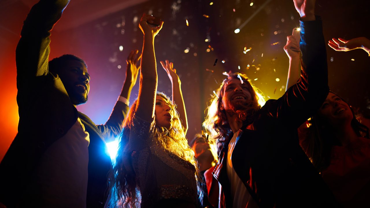Group of carefree excited people dancing under falling confetti and waving hands in air at musical concert in nightclub